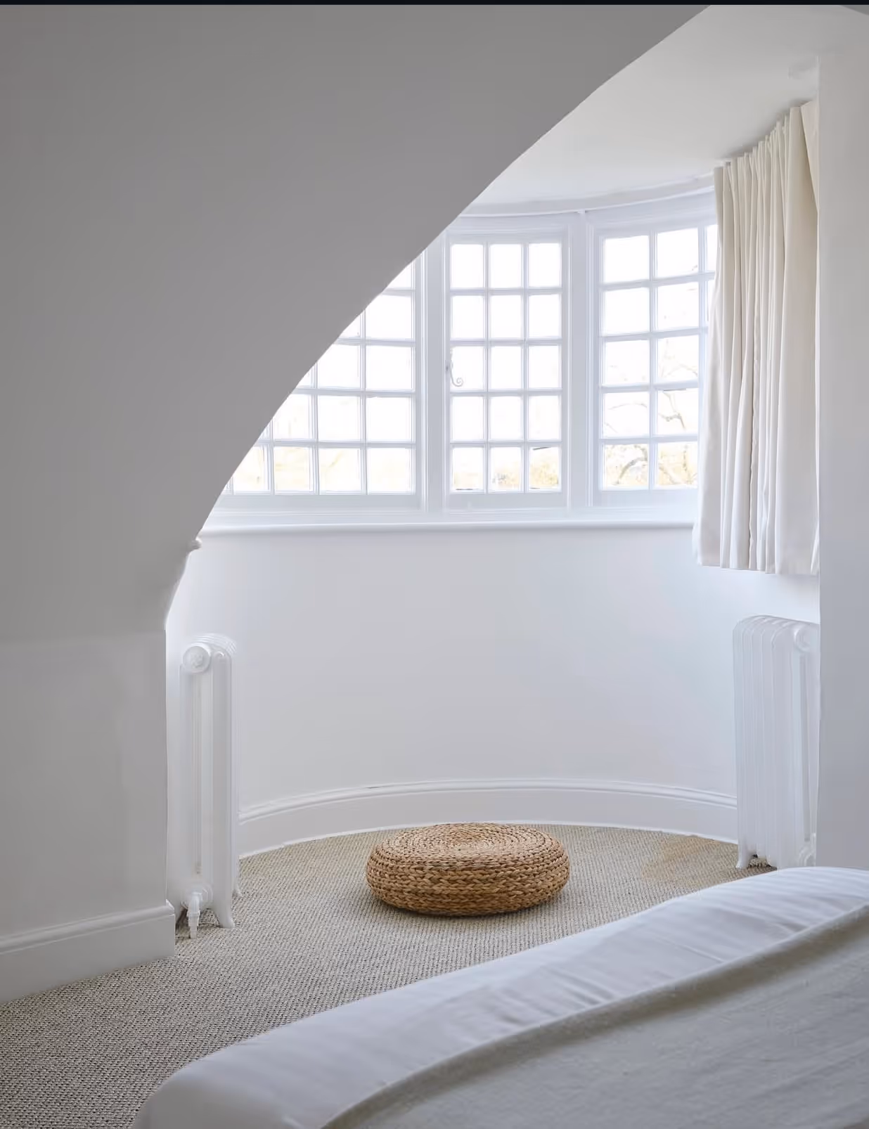 White attic bedroom with a curved bay window, a jute pouf on sisal carpet and a bed in the foreground.