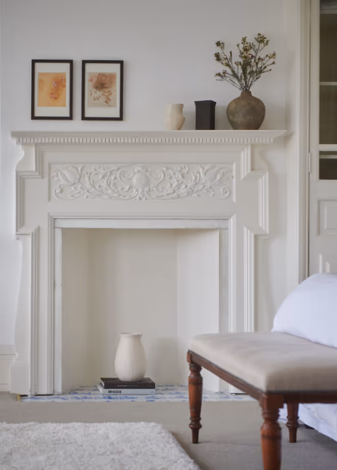 Ornate white fireplace mantel in a bedroom, with framed prints and a dried-flower arrangement.