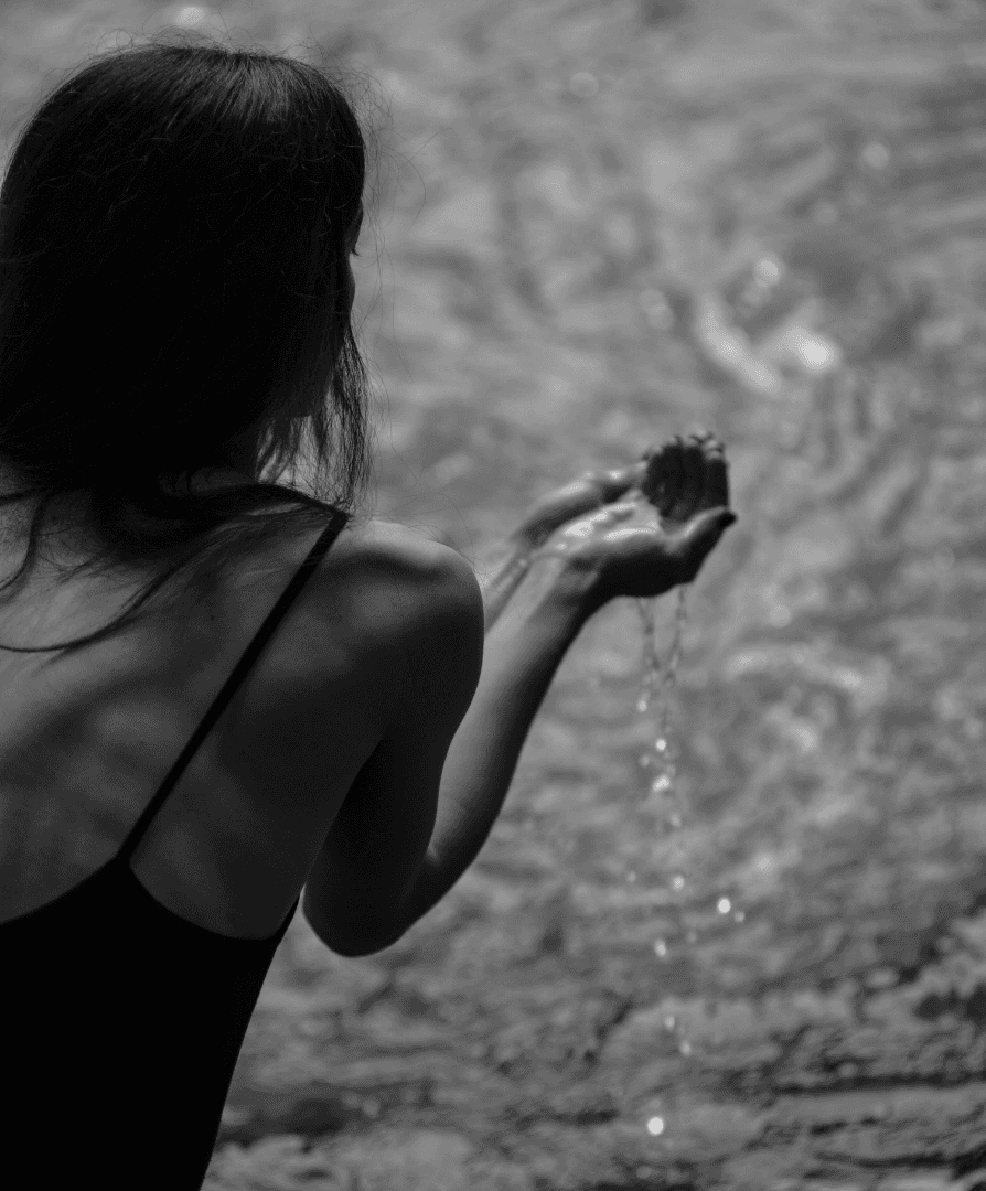 Black-and-white photo from behind of a woman cupping river water in her hands.