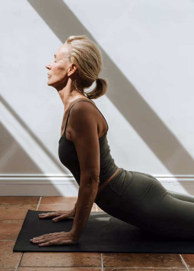 Older woman doing an upward-facing yoga pose on a mat, with striped sunlight on the wall behind her.