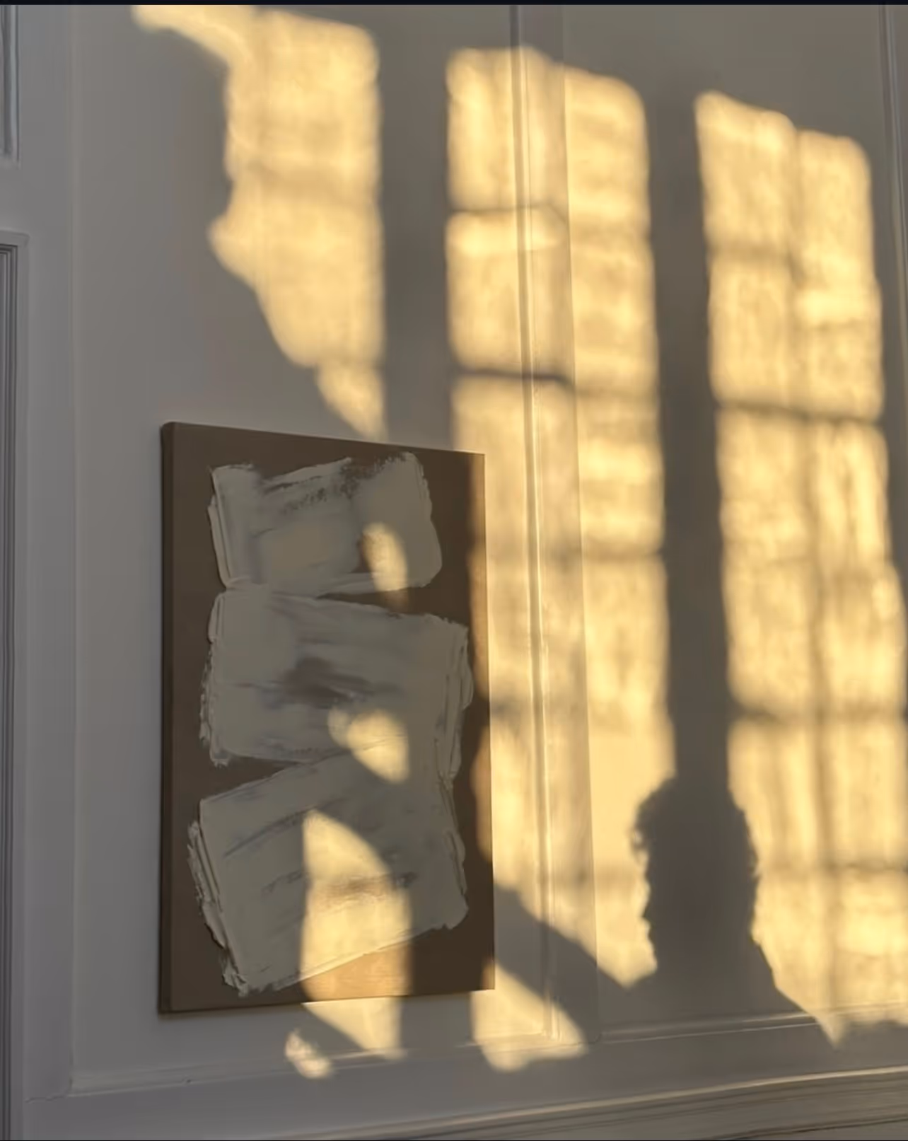 A textured abstract canvas on a wall with golden window-grid shadows and a person's silhouette.