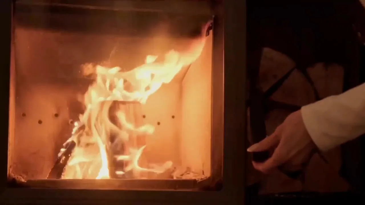 Flames in a glass-fronted wood burner, a hand reaching to the logs